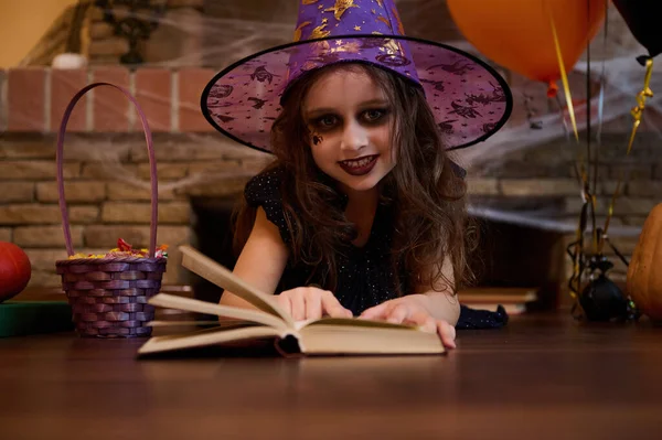 Mysterious little girl witch in wizards hat, looking at camera while learning sorcery, reading a spell book, lying on the floor against a spider web covered fireplace, surrounded by Halloween treats