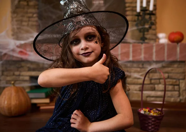 Close-up portrait of a gothic little child girl, with a spider painted on her cheek, wearing a wizards hat and witch dress, shows thumb up, looking at camera. Halloween party. Enchantress. Sorceress