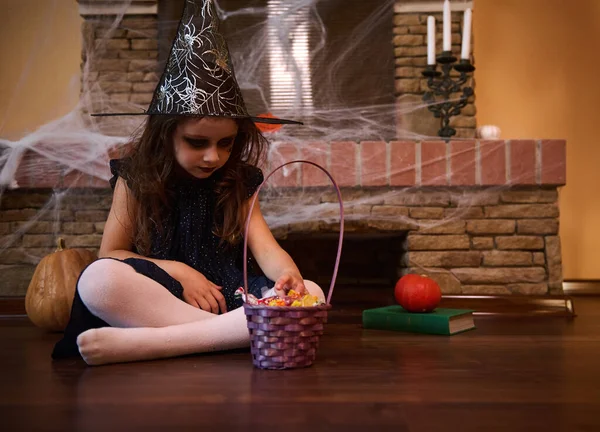 A gothic little child girl looking like a witch, wearing black dress and wizards hat, sitting by a cobweb covered fireplace, surrounded by Halloween decoration at home. Halloween trick and treat.