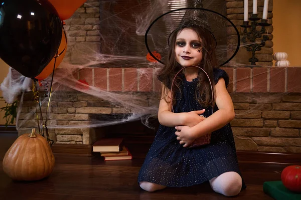 Pretty little girl, sorceress in wizards hat and smokey eye makeup, hugging basket of Halloween treats, sitting on floor surrounded by spell books and Jack-O-Lantern, against a fireplace with cobweb