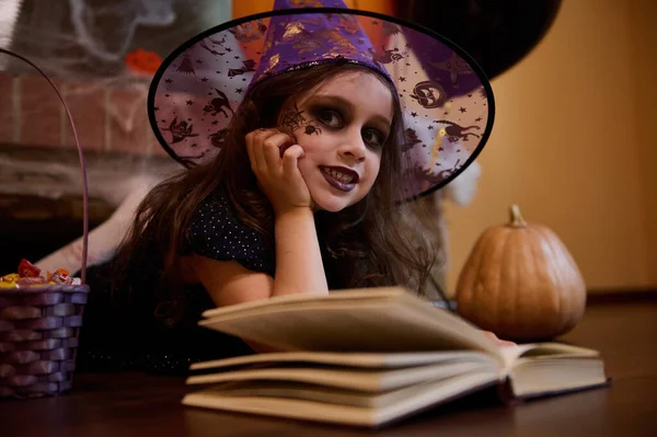 Inspired little girl dressed as a witch in a wizards hat, cutely smiles looking mysteriously at camera while reading a sorcery spell book, lying on the floor against a spider web covered fireplace