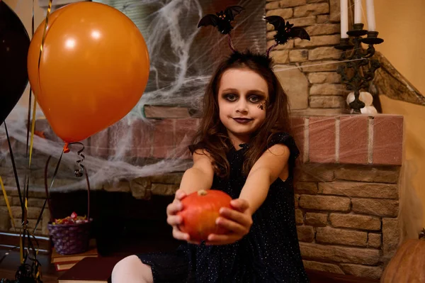 Caucasian gothic girl enchantress with black spider paint on her cheek and bat hoop, holds out a bright orange pumpkin, sits by a cobweb-covered fireplace and looks at the camera. Halloween home decor