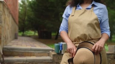 Sabit tabanlı: Latin Amerikalı güzel kadın çiftçi, başarılı tarımcı, bej önlüklü, hasır şapkalı ve uzağa bakıyor, bir kır evinin arka bahçesinde duruyor.