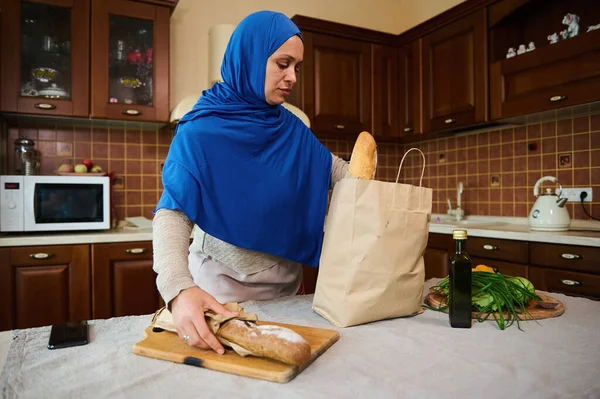Middle-Eastern Muslim woman with head covered in blue hijab, removes baguettes of wholegrain bread from a paper packet while unpacking grocery shopping bag. Food delivery. Healthy eating