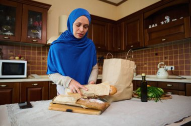 Müslüman Ortadoğulu güzel kadın ev hanımı, mavi tesettürlü, tahıllı ekşi ekmeği ahşap bir tahtaya yerleştirirken, ev mutfağındaki poşetteki taze yiyecekleri boşaltıyor.