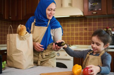 Mavi tesettürlü mutlu Orta Doğulu Müslüman kadın ev hanımı, sevgili bir anne ve alışveriş sonrası bavullarını açan sevgili kızı. Çevrimiçi süpermarketlerin müşterileri. Yemek teslimatı