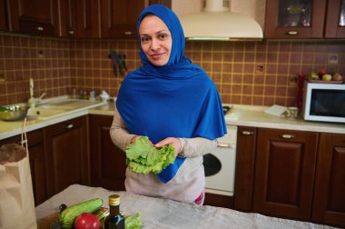 Büyüleyici Orta Doğulu Müslüman kadın mavi başörtüsü takıyor, elinde taze organik salata yapraklarıyla mutfak adasının yanında duruyor, taze çiğ vegan salatası pişiriyor, kameraya bakıp gülümsüyor.