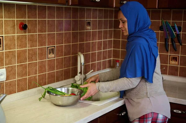 Arab Muslim woman with head covered in a blue hijab, stands by a sink in the kitchen at home, washes fresh organic vegetables and salad leaves under running water in a tap. Cooking.