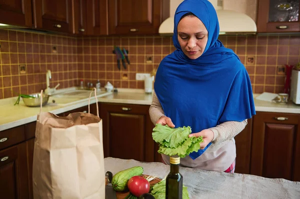 Arab Muslim woman with head covered in blue hijab, holding leaves of fresh salad, standing by a kitchen island, unpacking eco paper bag with food and groceries when arriving from grocery shopping