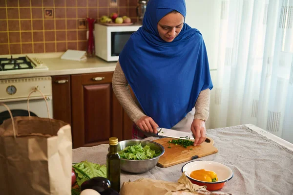 Pleasant Middle-Eastern Muslim woman housewife, with her head covered in a blue hijab, chopping greens on a wooden board, cooking delicious healthy meal