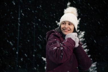 Sıcak yün şapkalı ve eldivenli gülümseyen çok ırklı bir kadının portresi. Karlı bir akşamda bir şehir caddesinde. Kar yağışı. Mutlu Noeller ve kış tatili konsepti
