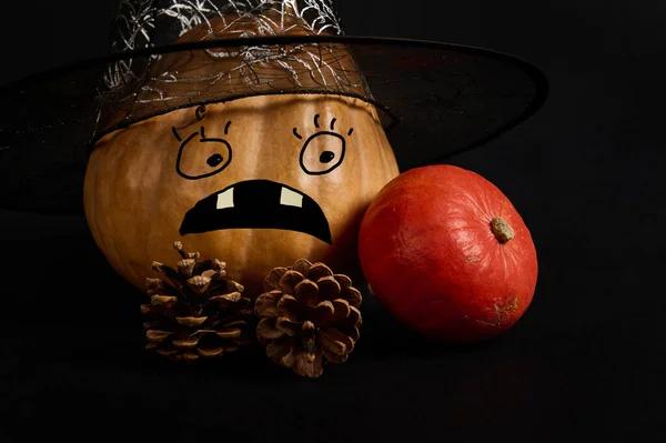 A whole orange pumpkin in wizard hat with a scary painted face and pine cones, on black background with copy advertising space. Jack-O-Lantern. Halloween trick and treat. Autumnal festival. Harvesting