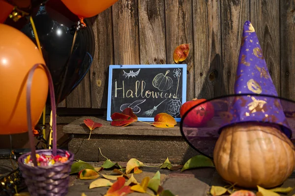 A wooden threshold or house porch, decorated for Halloween party with orange black air balloons, whole pumpkin in purple wizards hat, fallen dry colorful autumn leaves and a violet basket with sweets