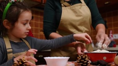 Kafkasyalı anaokulu çocuğu, sevimli küçük kız, sevimli kız elf çemberi ve bej şef önlüğü takmış, hamurlu bir kaseye şekerli meyve eklerken, annesi mutfakta noel pastası hamuru yoğuruyor.