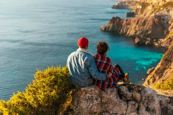 海沿いの崖の端に男と女が腰を下ろしている 海のそばの夕日に恋をしているカップル リアビュー 男と女が旅をしている スタイリッシュなカップルが海岸の夕日を満たしています — ストック写真