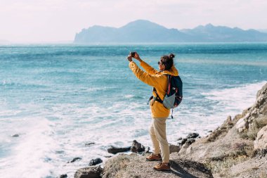 Arka planda deniz manzaralı, telefonu olan bir gezgin. Sırt çantalı sarı ceketli bir turist kız elinde bir telefon tutuyor. Seyahat ve aktif yaşam tarzı konsepti. Boşluğu kopyala