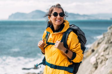 Turist kıyafetli genç bir kadın denizin arka planında duruyor ve kameraya bakıyor. Sırt çantalı gezgin bir kadın kameraya gülümsüyor. Gülümseyen bir turist portresi. Gülümseyen turist