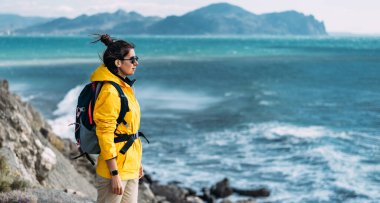 Sırt çantalı bir turist denize karşı, panorama. Denize karşı turist kıyafeti giymiş bir kadının portresi. Seyahat ve aktif yaşam tarzı konsepti. Sarı ceketli bir gezginin portresi. Boşluğu kopyala