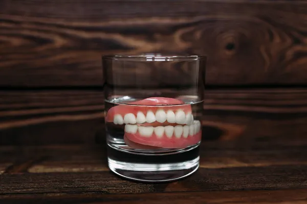コップ一杯の水の中の入れ歯 歯列矯正ケア 顎の完全な取り外し可能なプラスチック製の入れ歯 アクリル製の入れ歯2つ 上顎と下顎に偽の歯がある 歯や偽の歯 クローズアップ — ストック写真