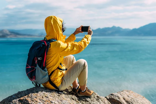 リアビューの背景に携帯電話を持つ旅行者 黄色のジャケットを着た観光客の女の子は 彼女の手に携帯電話を持っています クローズアップ 観光客が携帯電話で海の写真を撮る — ストック写真