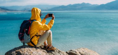 Denizin arka planında bir kız gezgin. Macera, seyahat ve yürüyüş konsepti. Denizin arka planında telefonu olan bir gezgin, panorama. Deniz kenarında turist kıyafeti giymiş bir adam.