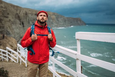 Denizin arka planında sırt çantası olan sakallı bir gezgin. Kırmızı elbiseli ve güneş gözlüklü bir gezginin portresi. Sırt çantalı bir turist denizin arka planında duruyor. Boşluğu kopyala