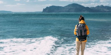 Arka planda deniz manzaralı sırt çantalı bir gezgin. Mavi denizin arka planında bir kadın gezgin, panorama. Deniz kıyısında yalnız bir gezgin. Turist bayrağı. Boşluğu kopyala