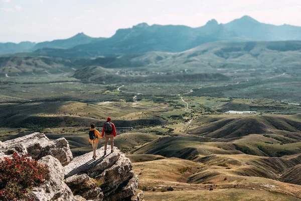 観光機器の男と女が岩の上に立っていて パノラマビューを賞賛しています バックパック付きの2人の観光客は山の頂上からの景色を楽しむ バックパッキング旅行 スペースのコピー — ストック写真