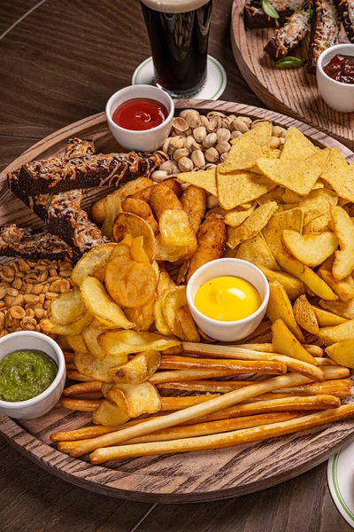 Set of assorted beer snacks and sauces served in a wooden box on dark concrete background. Potato chips, nachos, french fries, rye bread croutons, sausages, spring rolls, cheese, herring toasts