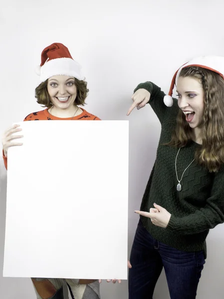 Twee mooie jonge vrouwen dragen santa hoeden en bedrijf leeg teken voor kopiëren ruimte. — Stockfoto