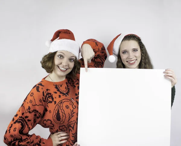 Twee mooie jonge vrouwen dragen santa hoeden en bedrijf leeg teken voor kopiëren ruimte. — Stockfoto