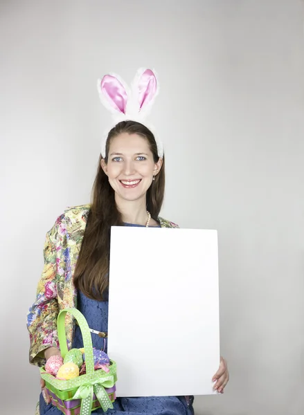 Bella giovane donna che indossa giacca colorata, abito blu e orecchie di coniglio in possesso di cesto di Pasqua e segno bianco — Foto Stock