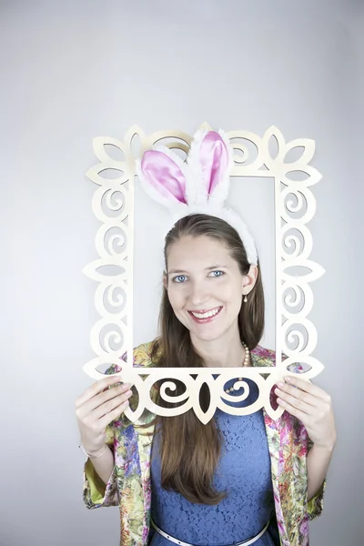 Pretty young woman wearing colorful jacket, blue dress and rabbit ears coming out of wooden picture frame — Stock Photo, Image