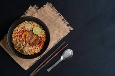 yummy instant noodles or ramen noodles with tuna fish and lemon slice in a classic black bowl with a stainless spoon and wooden chopsticks, still life Thai food top view on a dark kitchen table