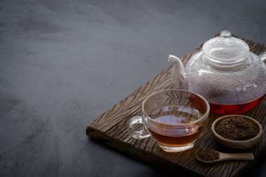 Hot tea in a glass teapot and cup decorated with dried tea leaves in a little wooden bowl and mini spoon on the old wood plank with dark kitchen table background