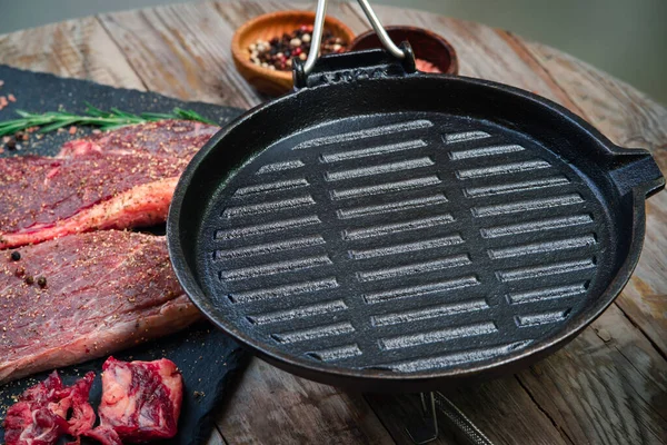 empty cast iron pan is seasoned with oil for dinner preparation with raw beef steak rosemary leaf herbs garlic, and spices black pepper on a wooden table background, pan seared cooking, grill meat