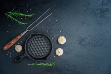Empty cast iron pan and bbq steak fork on dark gray cooking background decoration with rosemary garlic and peppercorn, top view with copy space for your text.