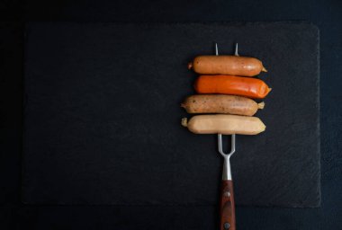 Çeşitli türde çiğ barbekü sosisleri aperatifleri yapmaya hazır bir tahta çatalla çarpıtılıyor. Üst arkaplanda yemek pişirmek için gereken malzeme. Yemek menüsü için yer var.