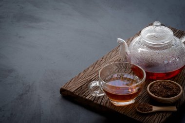 Hot tea in a glass teapot and cup decorated with dried tea leaves in a little wooden bowl and mini spoon on the old wood plank with dark kitchen table background