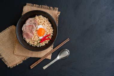 instant noodles or ramen noodles with boiled egg and slice beef and big red chillies in a classic black bowl with a stainless spoon and wooden chopsticks, Thai food top view on a dark kitchen table