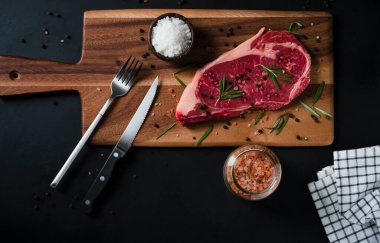 Himalaya Pembe tuzu, biberiye yaprağı otları ve ahşap kesme tahtasındaki baharatlı karabiberli çiğ biftek. Metnin için kopyalama alanı olan koyu arkaplan üzerindeki üst görünüm