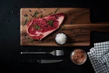 Himalaya Pembe tuzu, biberiye yaprağı otları ve ahşap kesme tahtasındaki baharatlı karabiberli çiğ biftek. Metnin için kopyalama alanı olan koyu arkaplan üzerindeki üst görünüm