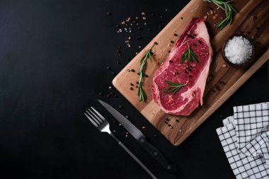 Himalaya Pembe tuzu, biberiye yaprağı otları ve ahşap kesme tahtasındaki baharatlı karabiberli çiğ biftek. Metnin için kopyalama alanı olan koyu arkaplan üzerindeki üst görünüm