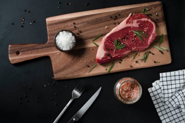 Himalaya Pembe tuzu, biberiye yaprağı otları ve ahşap kesme tahtasındaki baharatlı karabiberli çiğ biftek. Metnin için kopyalama alanı olan koyu arkaplan üzerindeki üst görünüm