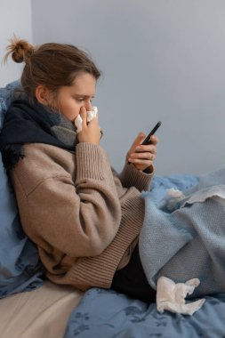 Beyaz kazaklı, siyah eşarplı, burnu kağıt peçeteye sümküren hoş beyaz bir kadın. Kız yatakta oturuyor ve internetten ilaç dağıtımını arıyor. İnternetten ilaç alımı.
