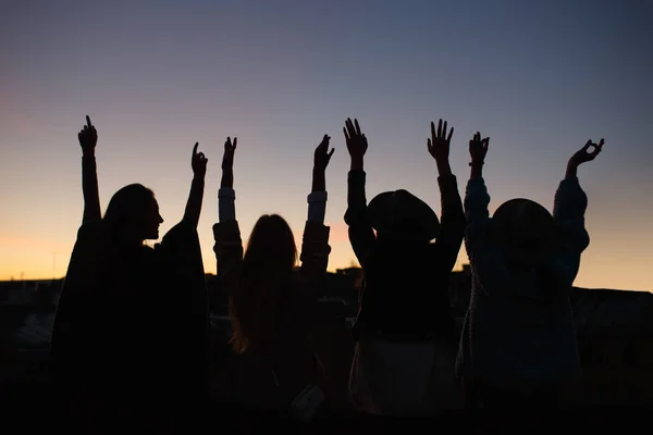 Silhouette photo of four attractive happy young women sitting with their backs and hands up. Hipster girls enjoy life. Few females in hats. Sunset on background. Freedom concept. Recreation leisure.