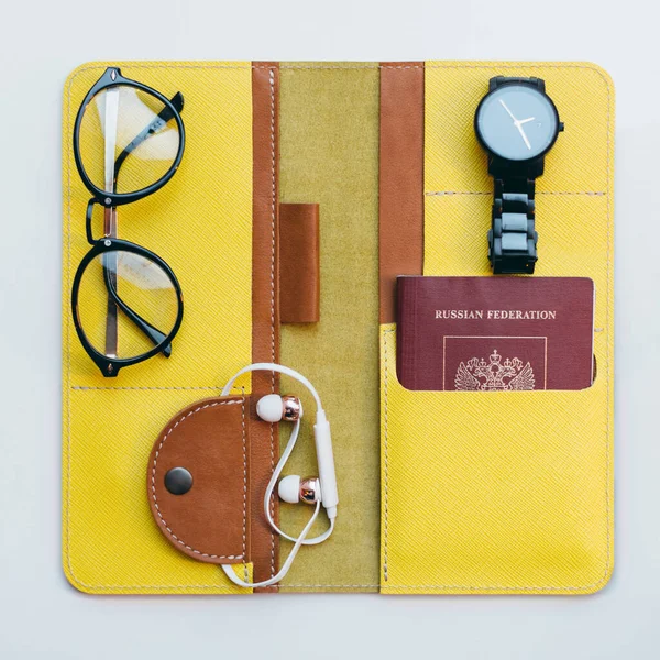 Bright yellow opened travel wallet for documents. Inside passport or id card, boarding pass and money. Near black glasses, hand watches and white headphones. Flat lay. Vacation and tourism concept.