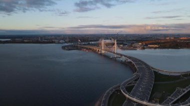 Neva nehri boyunca uzanan kıvrımlı çok şeritli otoyolun havadan görünüşü. Bir sürü araba yolda hızlı hareket ediyor. Arka planda Pylon köprüsü ve Zenit Arena stadyumu var. Kamera geri gidiyor.