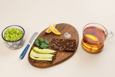 Avokado tostu için malzeme içeren ahşap tahta. Avokado dilimli koyu çavdar ekmeği, taze fesleğen yaprağı, limon dilimleri ve susam tohumları. Kase ve çayın içinde guacamole 'nin yanında. Sağlıklı kahvaltı kavramı
