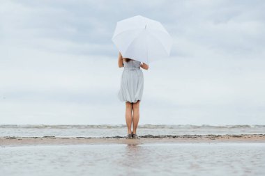 Gri elbiseli, beyaz bir şemsiyenin altında sırtı kameraya dönük duran ve denize hayran olan tanınmayacak kadar güzel bir kız. Barış ve rahatlama kavramı. Seçici odaklanma. Boşluğu kopyala.